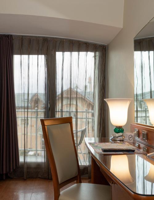 Doppelzimmer mit großzügigen, weißen Betten und Schreibtisch, mit einem cremefarbenen Sessel sowie mit Blick auf bodentiefe Fenster mit Balkon und einer Aussicht auf das Villenviertel Blasewitz in Dresden